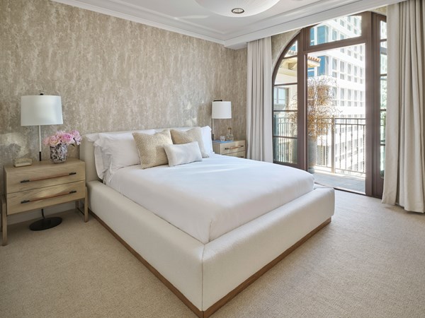 Maybourne Residence 3 bedroom: A white double bed sits in the foreground. To the left sits a wooden bedside table with pink flowers. A large window with curtains is to the right.