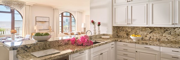 Maybourne Beverly Hills Residence Kitchen: A white kitchen with marble countertops is in the foreground. To the right is a white bowl with lemons. There are pink flowers in the centre. To the left there is a bowl of watermelons on the counter and underneath is a wine cooler.