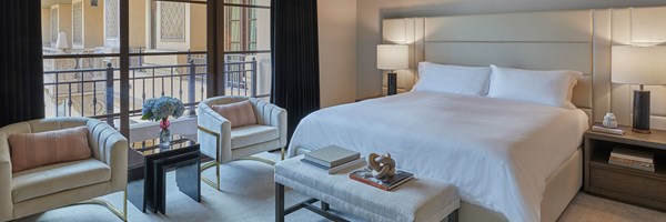 Maybourne Beverly Hills Residence Bedroom: A white double bed sits in the foreground. A wooden bedside table with books and a lamp sits to the right. To the left sits two white armchairs with pink cushions. In the left background there is a large window.