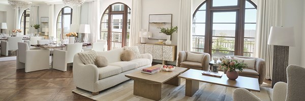 Maybourne Beverly Hills Residence Living Room: A white couch and wooden coffee table sit in the foreground. A large rug sits underneath. Table and chairs sit to the left. Two large window sit in the background.