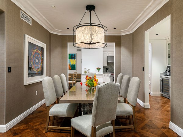 Maybourne Beverly Hills Residence Dining Area: In the foreground there is a wooden dining table with 8 chairs around it. Red and yellow flowers sit on the table. A chandelier light is hung from the ceiling. To the left is a picture frame.