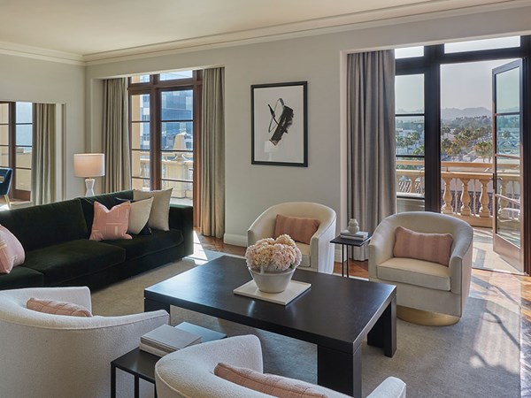 Maybourne Beverly Hills Residence: A brown wooden coffee table and pink flowers is in the foreground. Four white armchairs with pink cushions surround the table. To the left is a green couch with cushions. Two large windows sit in the background