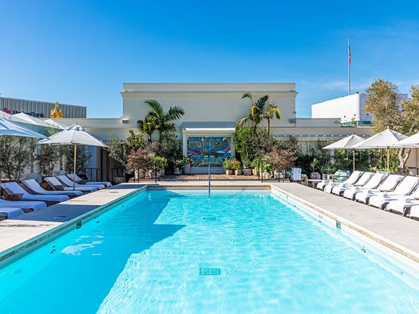 Maybourne Beverly Hills Residence Swimming Pool: A large swimming pool sits in the foreground. To the left and right there are white sun loungers and umbrellas. In the background there is a mural and green trees and plants either side.